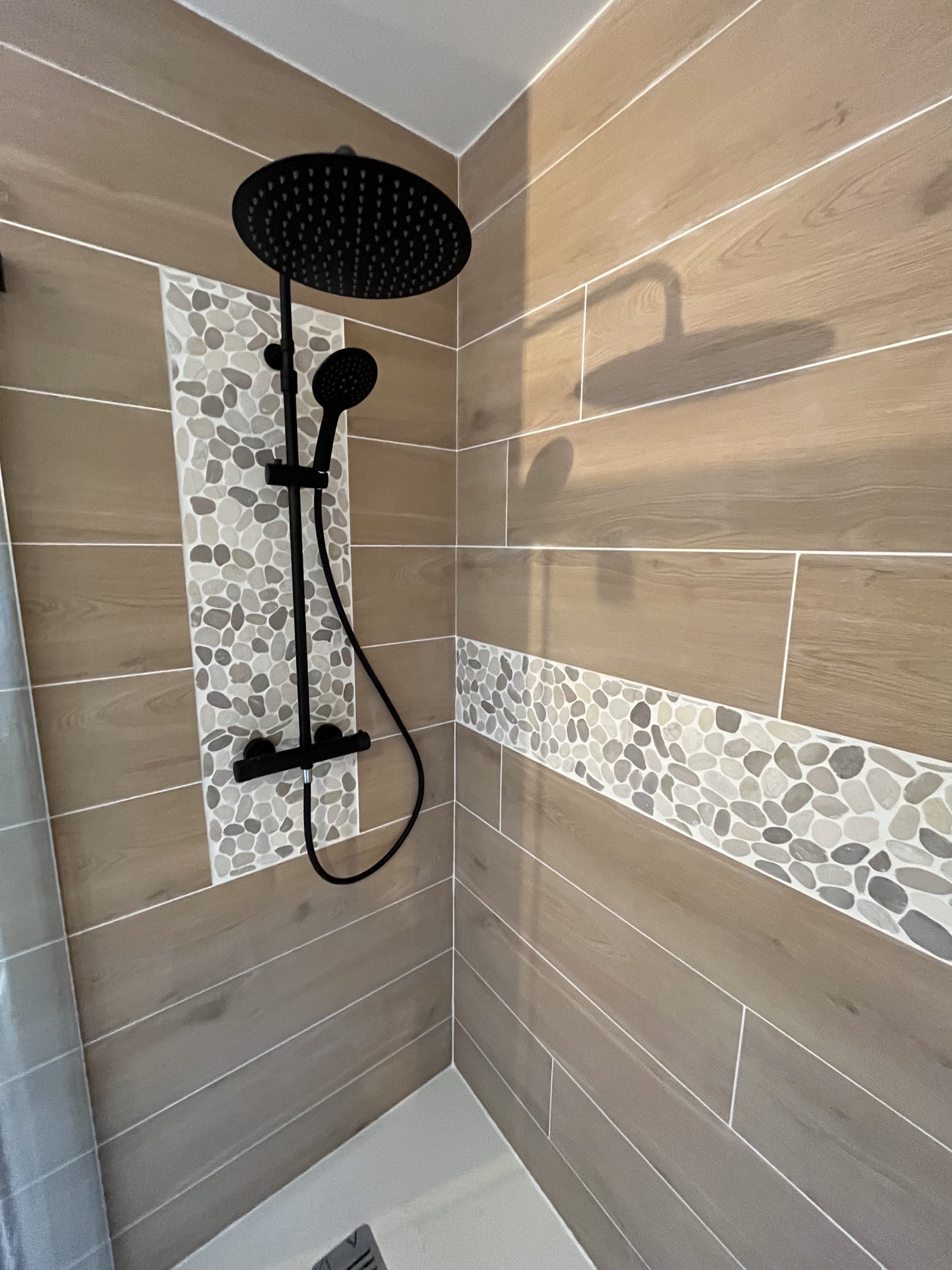 Détail d'une colonne de douche noire sur carrelage effet bois et galets